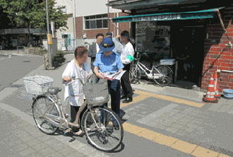 制服警察官が交番前で自転車盗難防止のためのパンフレットを配布している写真